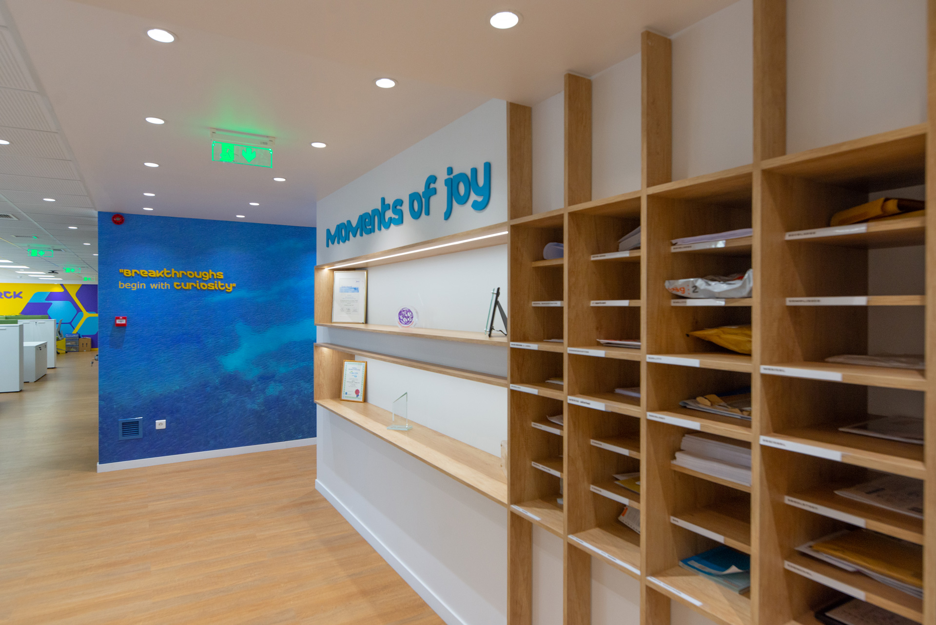 Merck Group workplace project by STIRIXIS Group showing lounge area with moments of joy brand wall and warm wood shelving unit