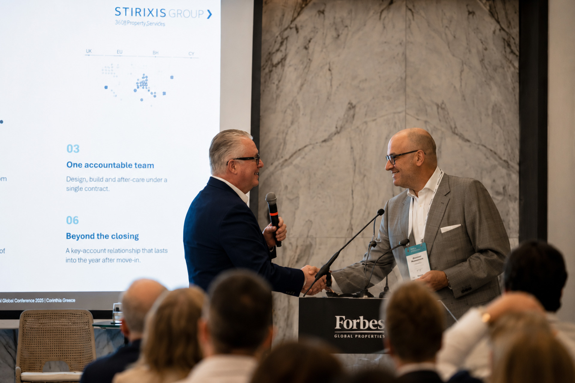 Alex Athanassoulas, President and CEO of STIRIXIS Group, shaking hands with Michael Jalbert, CEO of Forbes Global Properties, on stage at the Forbes Global Properties Annual Global Conference 2026, Corfu, with the STIRIXIS Group presentation visible on screen.